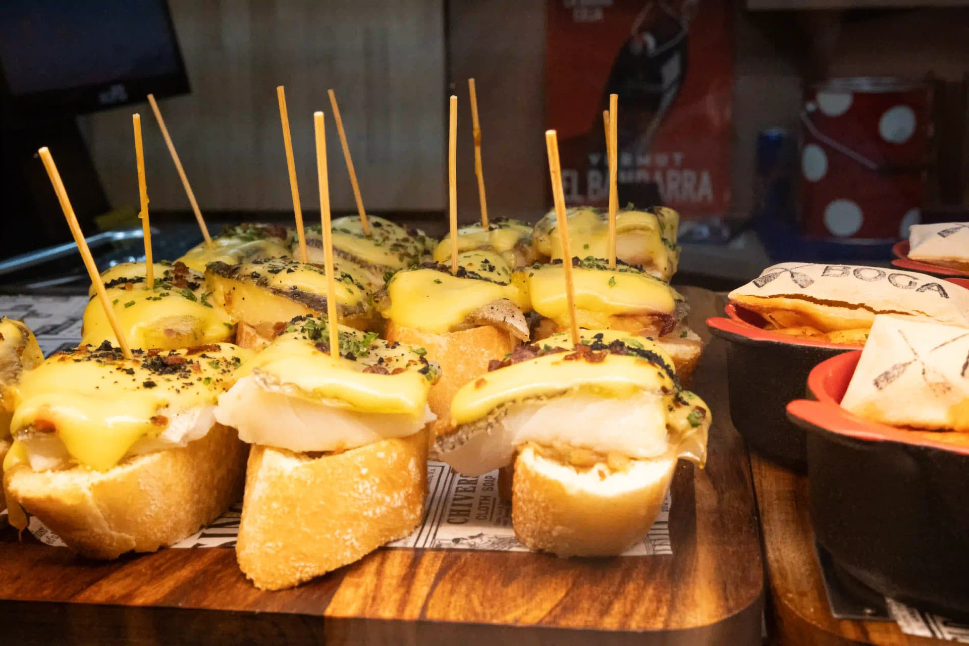 Tabla de pintxos de pan gratinados con salsa y sujetos con palillo, junto a cazuelas de barro en la barra
