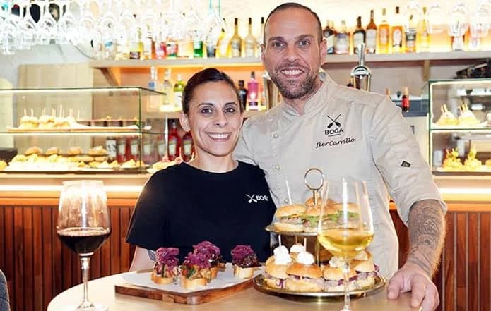 Eli e Iker Carrillo tras la barra de Boga sosteniendo dos bandejas de pintxos y dos copas de vino