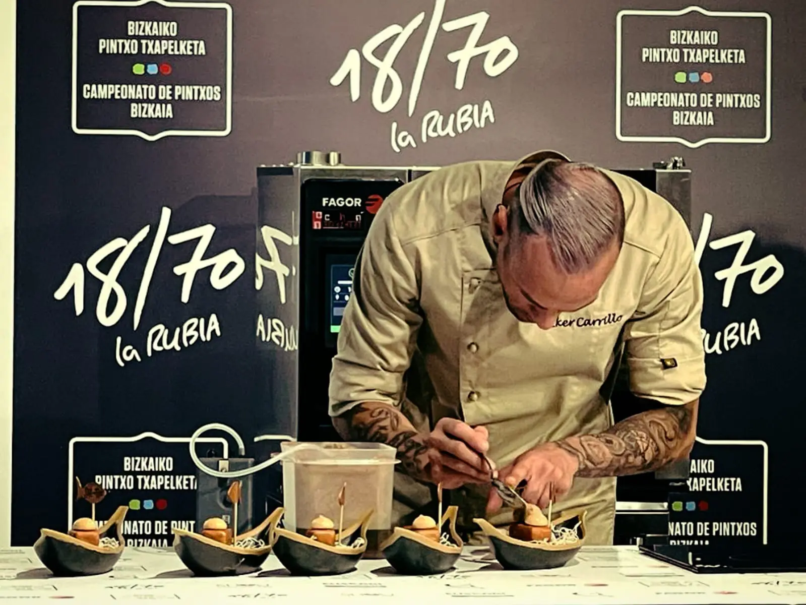 Pintxo gratinado sobre una baldosa de terrazo, rodeado de puntos de crema rosa, aceitunas negras y encurtidos