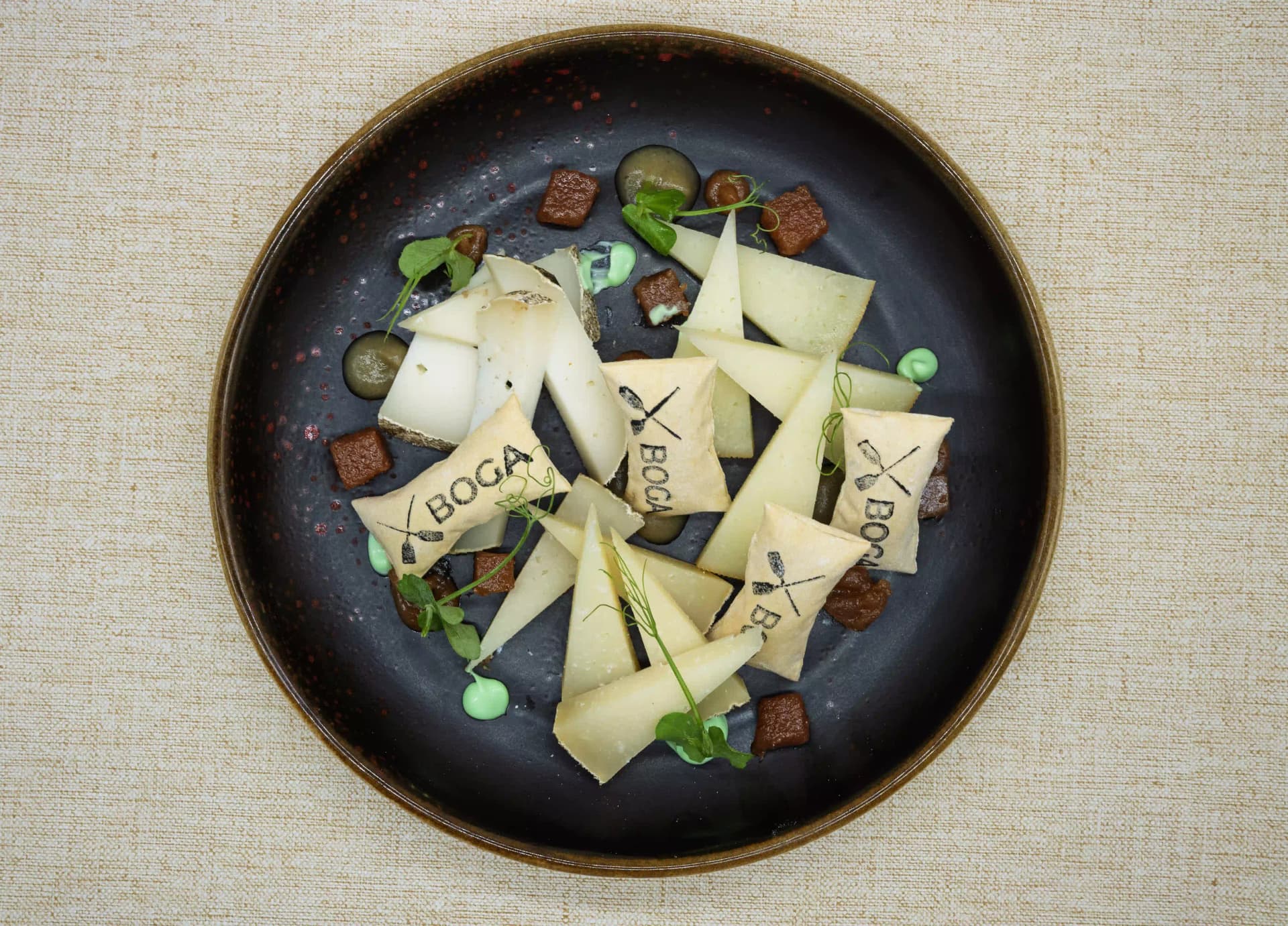 Láminas de queso de oveja con obleas serigrafiadas con el logo de Boga, puntos de crema y brotes, en plato negro