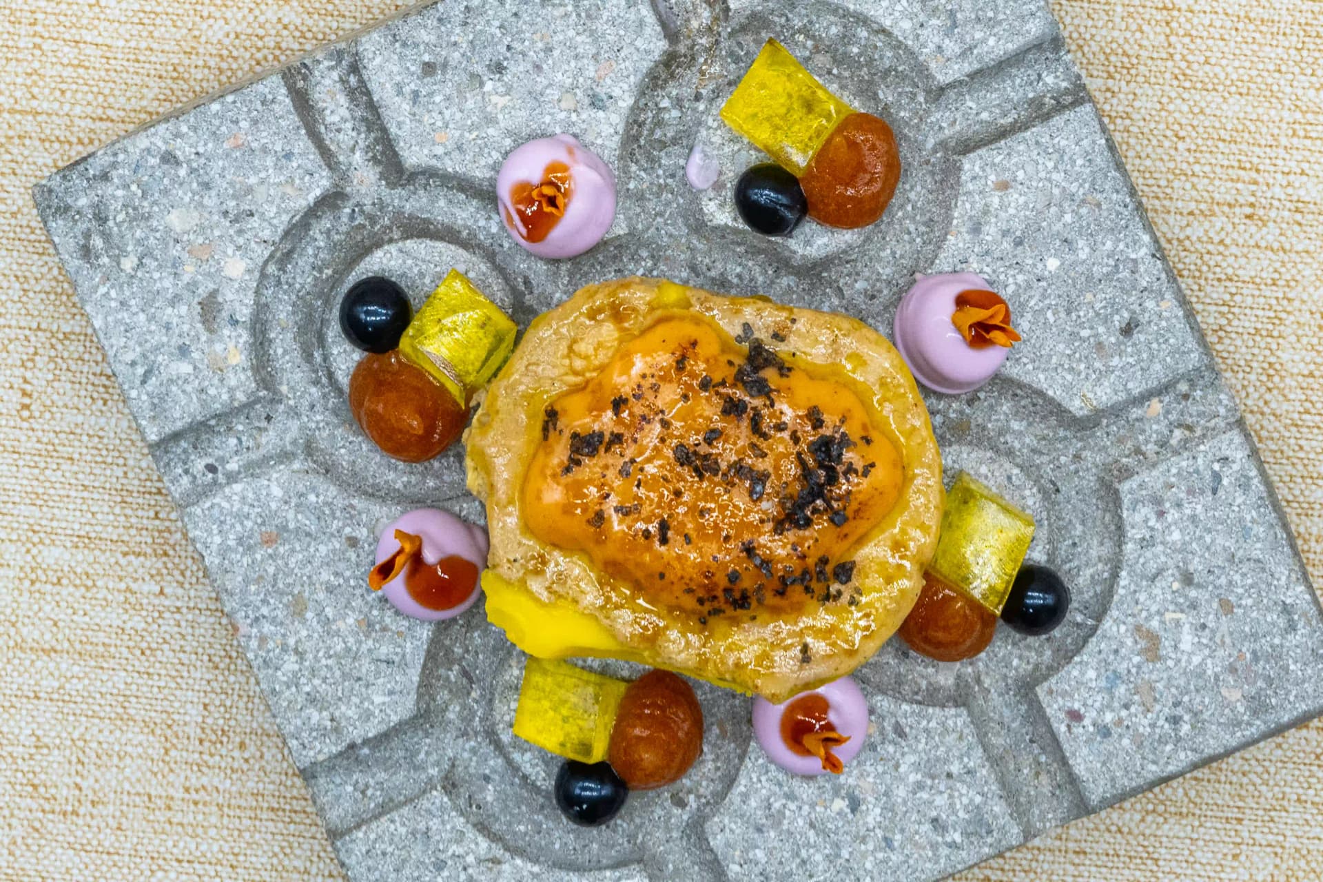 Pintxo gratinado sobre una baldosa de terrazo, rodeado de puntos de crema rosa, aceitunas negras y encurtidos